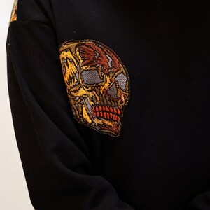 May include: Black long-sleeved sweater with a large embroidered skull design in shades of yellow, orange, and brown.