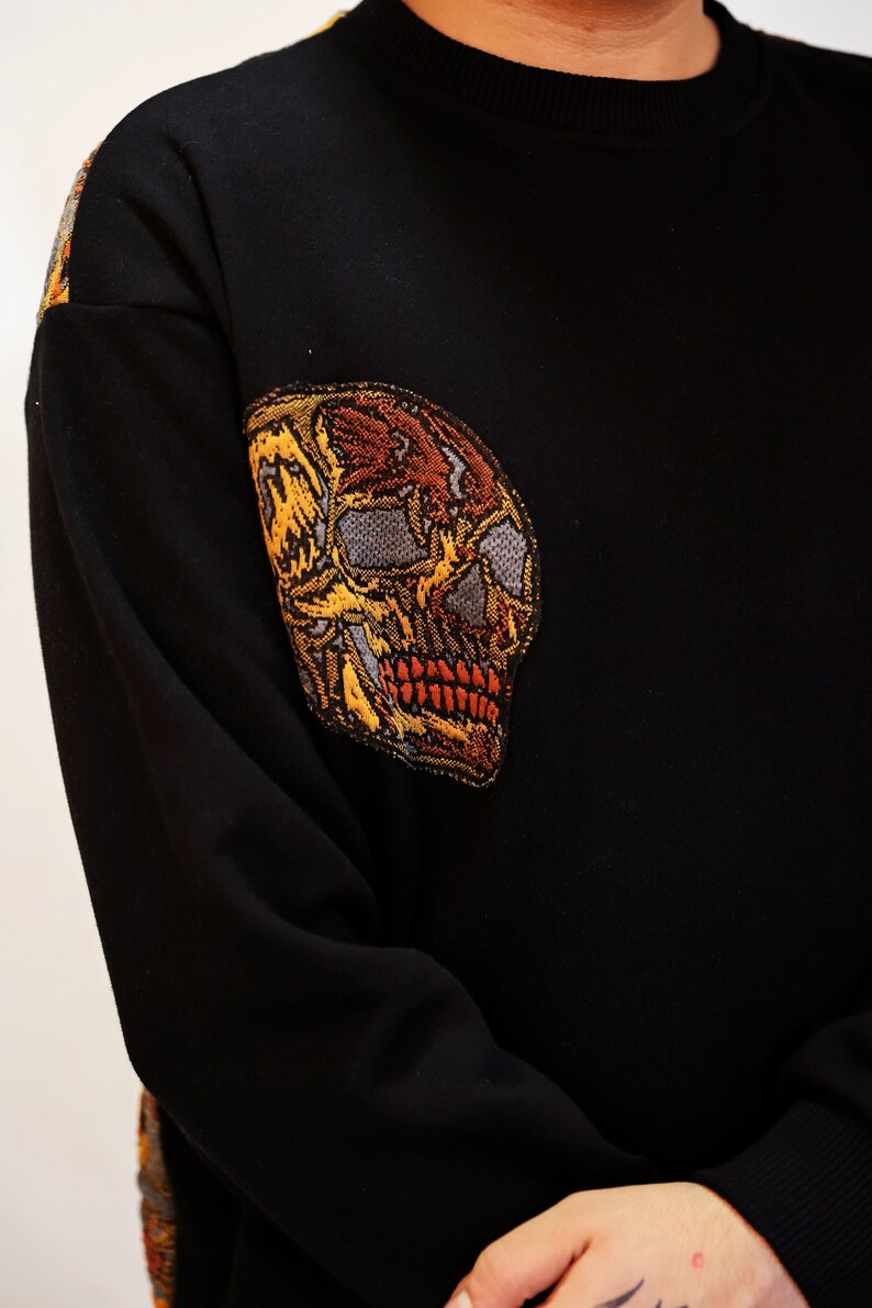May include: Black long-sleeved sweater with a large embroidered skull design in shades of yellow, orange, and brown.