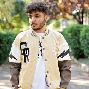 May include: A tan varsity jacket with brown leather sleeves. The jacket has a white lightning bolt design on the sleeve and a large letter "R" on the chest. The text "Life what happens when you busy making other plans" is printed on the jacket.