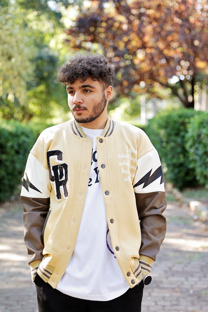 May include: A tan varsity jacket with brown leather sleeves. The jacket has a white lightning bolt design on the sleeve and a large letter "R" on the chest. The text "Life what happens when you busy making other plans" is printed on the jacket.