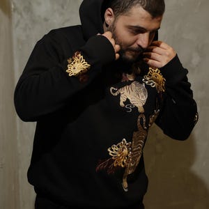 May include: Black hooded sweatshirt with embroidered tiger and floral designs in gold, brown, and cream colors. The sweatshirt features a drawstring hood and ribbed cuffs. The design includes a tiger and floral patterns on the front and sleeves.