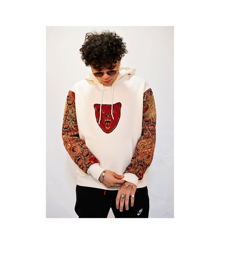 May include: A white hoodie with a red bear face embroidered on the front. The sleeves are patterned with a brown and red design.