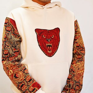 May include: A white hoodie with a red embroidered bear head on the front and a multi-colored embroidered pattern on the sleeves.
