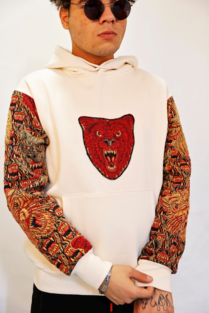 May include: A white hoodie with a red embroidered bear head on the front and a multi-colored embroidered pattern on the sleeves.