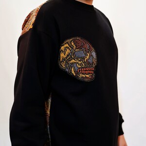 May include: Black sweatshirt with a colourful embroidered skull on the left side. The skull is detailed with red, yellow, and brown thread.