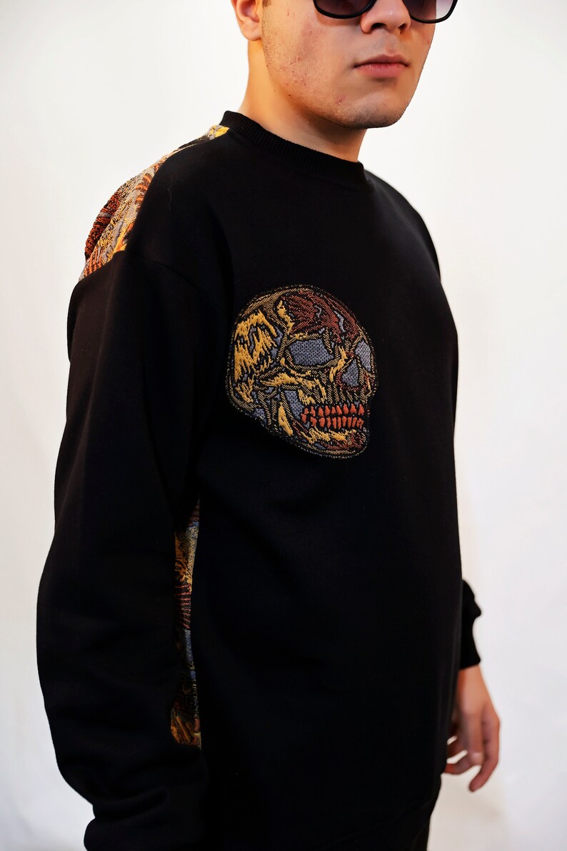 May include: Black sweatshirt with a colourful embroidered skull on the left side. The skull is detailed with red, yellow, and brown thread.