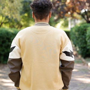 May include: A beige sweatshirt with brown and white sleeves. The sleeves have a black lightning bolt design. The sweatshirt has a ribbed collar and cuffs.