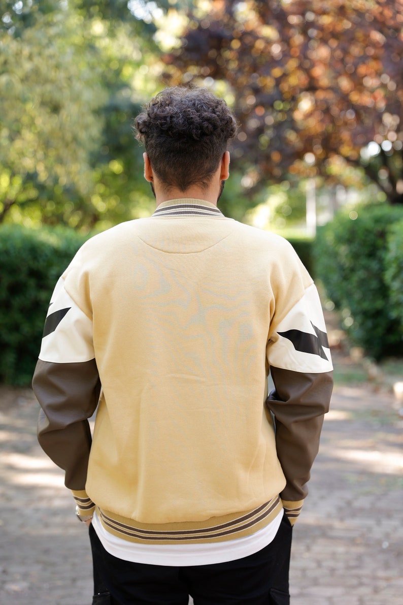 May include: A beige sweatshirt with brown and white sleeves. The sleeves have a black lightning bolt design. The sweatshirt has a ribbed collar and cuffs.