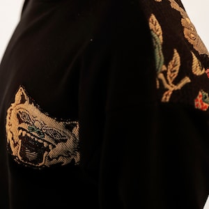 Puede incluir: Su&eacute;ter negro con un dise&ntilde;o bordado en dorado y marr&oacute;n en el hombro derecho y una cara de animal bordada en dorado y marr&oacute;n en la parte delantera.