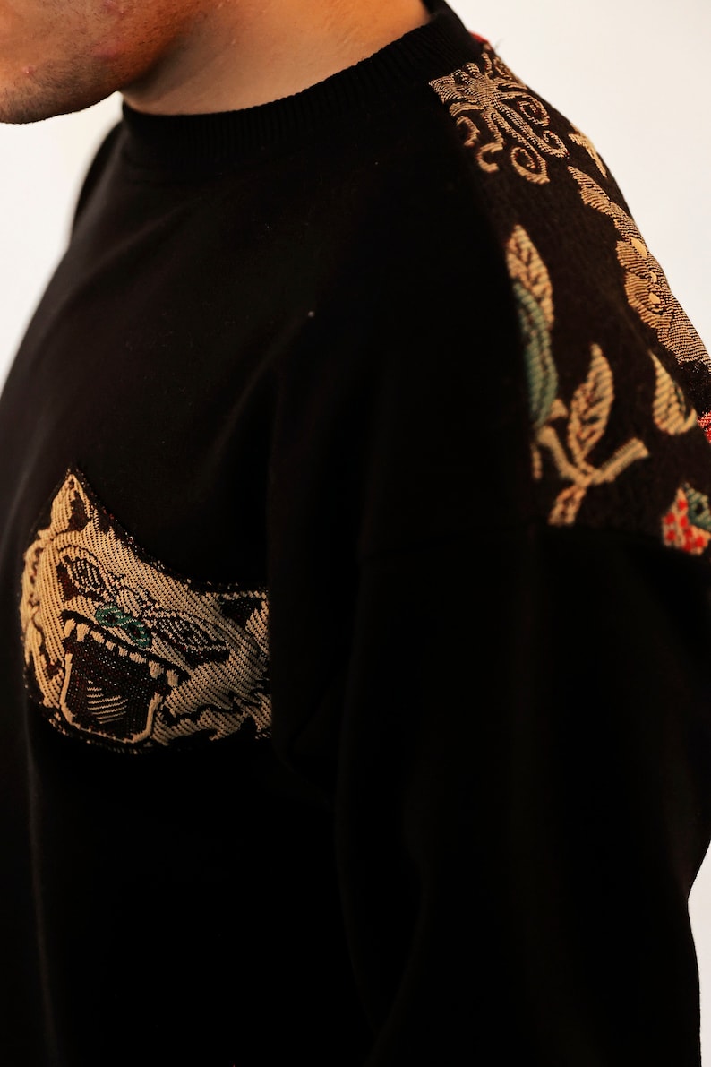 Puede incluir: Su&eacute;ter negro con un dise&ntilde;o bordado en dorado y marr&oacute;n en el hombro derecho y una cara de animal bordada en dorado y marr&oacute;n en la parte delantera.