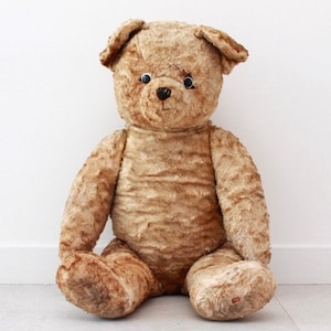 Peut inclure: Un grand ours en peluche brun avec des yeux en boutons noirs est assis sur un sol carrel&eacute; gris clair. L'ours a un aspect us&eacute; et vintage.