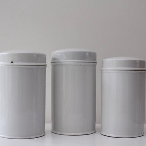 May include: Three white ceramic canisters with lids. The canisters are cylindrical and have a smooth, glossy finish.