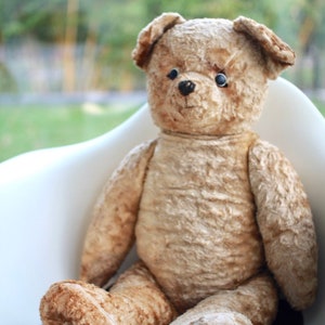 Peut inclure: Un grand ours en peluche brun avec des yeux noirs est assis sur une chaise blanche. L'ours a une texture us&eacute;e et duveteuse et est assis droit, les pattes reposant sur l'assise de la chaise.