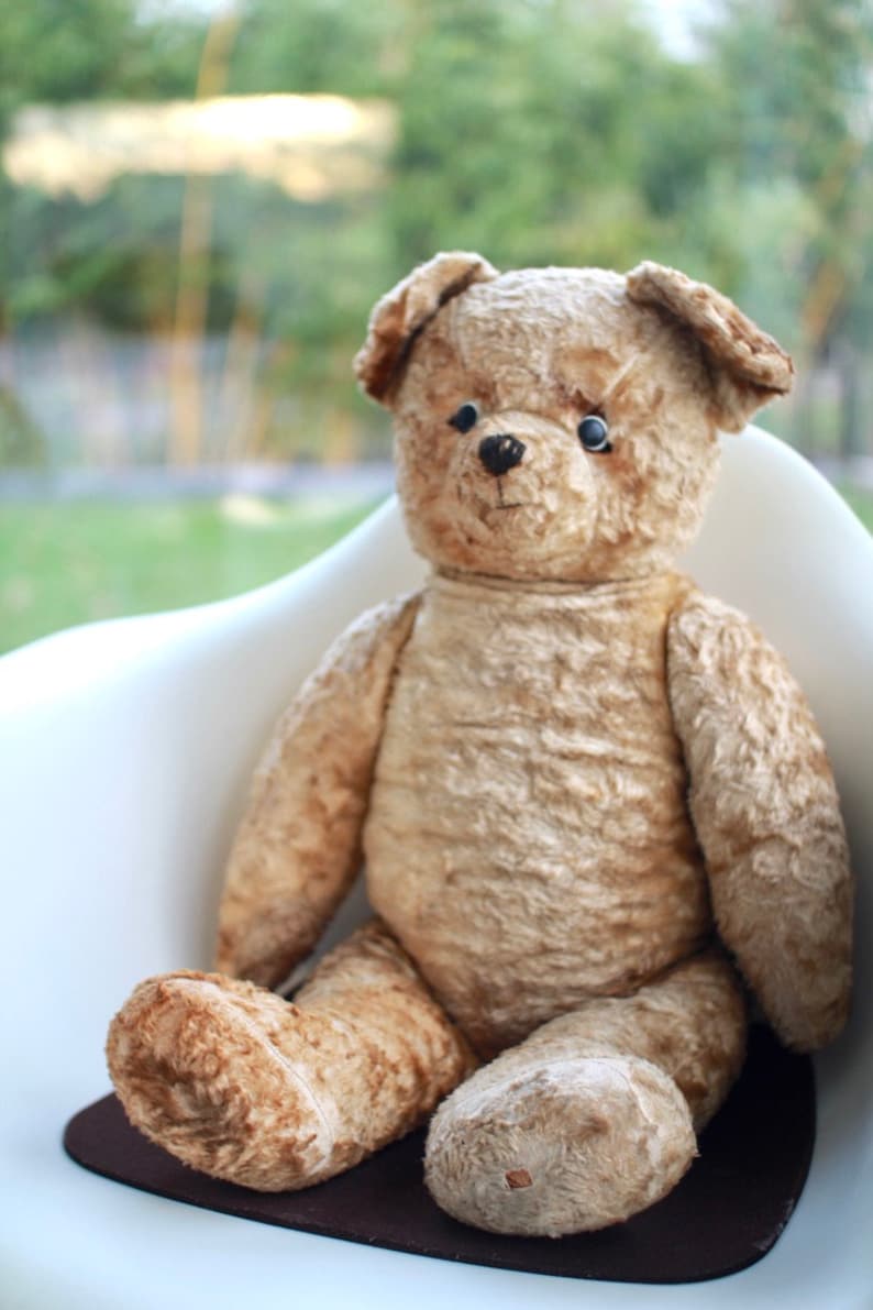 Peut inclure: Un grand ours en peluche brun avec des yeux noirs est assis sur une chaise blanche. L'ours a une texture us&eacute;e et duveteuse et est assis droit, les pattes reposant sur l'assise de la chaise.