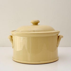 Puede incluir: Una cazuela de cerámica de color amarillo claro con tapa. El plato tiene dos asas y un pomo en la tapa. La cazuela es redonda y tiene un acabado brillante. Utensilios de cocina para hornear y servir.