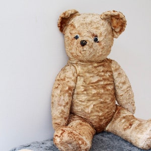 Peut inclure: Un ours en peluche vintage de couleur beige avec des yeux en boutons noirs et une texture us&eacute;e et duveteuse. L'ours est assis sur une surface duveteuse grise et blanche.