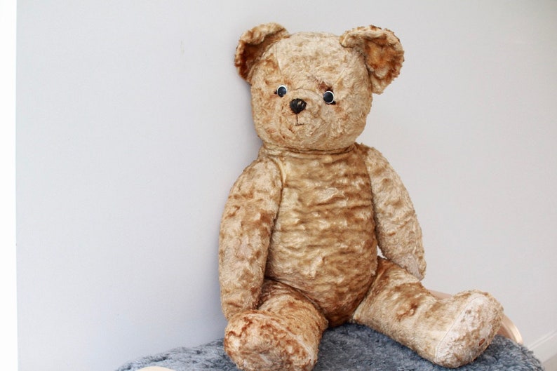 Peut inclure: Un ours en peluche vintage de couleur beige avec des yeux en boutons noirs et une texture us&eacute;e et duveteuse. L'ours est assis sur une surface duveteuse grise et blanche.