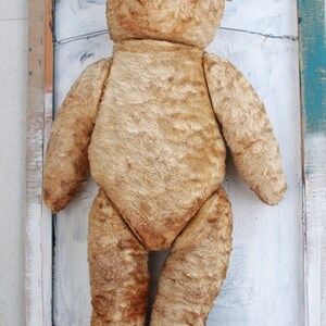 Peut inclure: Un ours en peluche vintage de couleur beige avec des yeux en boutons noirs. L'ours est debout devant un cadre en bois peint en blanc.