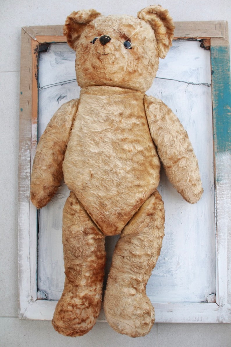 Peut inclure: Un ours en peluche vintage de couleur beige avec des yeux en boutons noirs. L'ours est debout devant un cadre en bois peint en blanc.