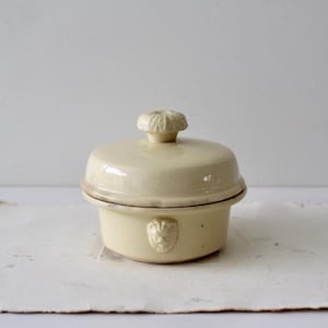 May include: A cream-colored ceramic lidded dish featuring a smooth, rounded body and a matching lid with a textured, flower-shaped knob. The exterior of the base is decorated with a small, molded lion head relief centered on the front side.