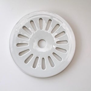 Może przedstawiać: Okrągły, biały ceramiczny filtr do zlewu z centralnym otworem i równomiernie rozmieszczonymi, podłużnymi otworami na obwodzie. Filtr ma błyszczące wykończenie i jest umieszczony na gładkim, białym tle.