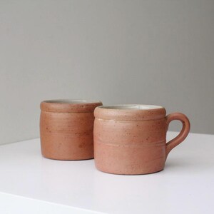 Pode incluir: Duas canecas de cerâmica rústicas de cor terracota. Uma caneca tem uma alça, enquanto a outra não tem. Ambas têm uma forma cilíndrica com uma borda ligeiramente alargada e um interior de cor clara. As canecas estão sobre uma superfície branca contra um fundo cinza neutro.