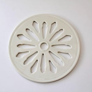 May include: A round, off-white ceramic trivet with a floral design. The trivet features a series of petal-shaped cutouts radiating from a central hole. The trivet is approximately 7 inches in diameter.