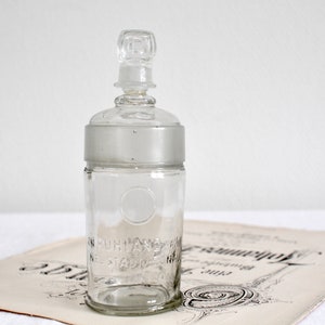 Op de afbeelding: Een heldere glazen fles met een mat glazen deksel en een glazen stop. De fles heeft een cirkelvormig reliëf en de tekst "The Standard" aan de zijkant.