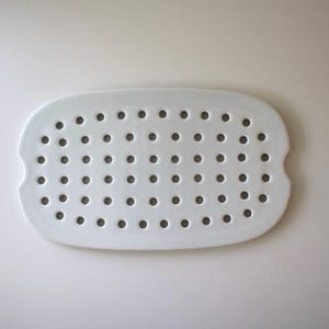 Może przedstawiać: Biały, owalny ceramiczny talerz z licznymi, równomiernie rozmieszczonymi okrągłymi perforacjami. Talerz ma lekko podniesioną krawędź i zaokrąglone rogi. Projekt sugeruje funkcjonalny przedmiot, prawdopodobnie do osuszania lub serwowania.