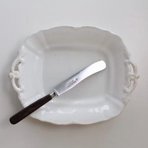 Könnte beinhalten: Eine weiße, rechteckige Servierschale mit gewellten Kanten und dekorativen Griffen. Ein Butterstreichmesser aus Edelstahl mit einem dunkelbraunen Griff liegt auf der Schale. Das Messer ist über der Schale angewinkelt.