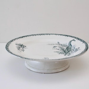 May include: White ceramic cake stand with a raised base. The plate features a teal and white bird and floral design around the rim and on the plate's surface. A decorative piece for serving desserts or displaying items.