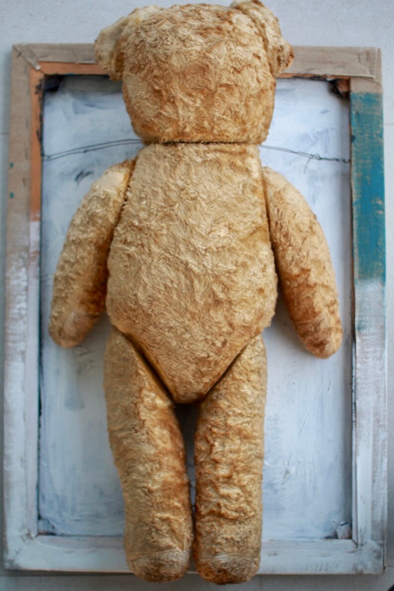 Peut inclure: Un ours en peluche vintage avec un pelage us&eacute; de couleur fauve. L'ours est debout dans un cadre en bois sur un fond blanc.