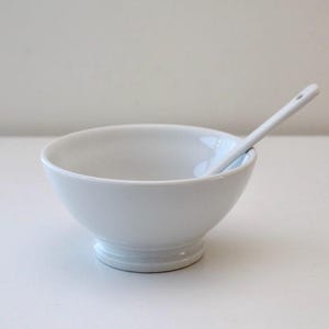 Könnte beinhalten: Eine weiße Keramikschüssel mit einem kleinen weißen Löffel darin. Die Schüssel hat eine abgerundete Form und einen leicht ausgestellten Rand.