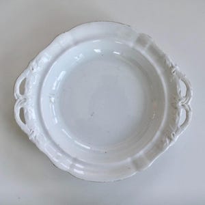 Könnte beinhalten: Eine weiße Servierplatte im antiken Stil mit gewelltem Rand und dekorativen Griffen. Die Platte hat eine leicht erhöhte Mitte und eine glänzende Oberfläche, ideal zum Präsentieren von Speisen oder als dekoratives Stück.