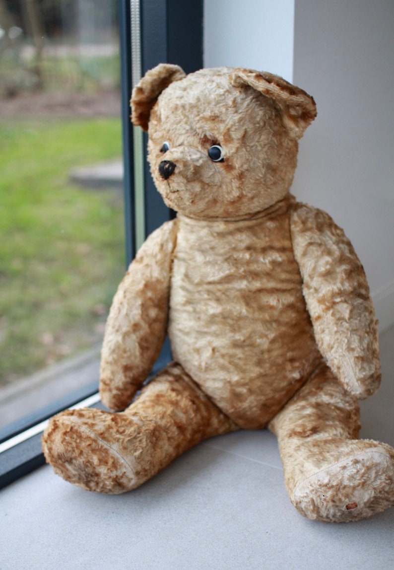 Peut inclure: Un grand ours en peluche brun clair avec des yeux noirs et un nez noir est assis sur un sol carrel&eacute; blanc. L'ours regarde vers la gauche de l'image.