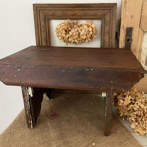 May include: A small, antique wooden step stool with a dark brown finish and distressed white paint on the legs. The stool has a rectangular top and is supported by four legs. The background includes a framed artwork and dried flowers.