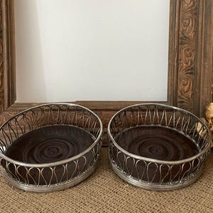 May include: Two silver-toned metal coasters with a dark brown wooden base. The coasters have a decorative wire design around the edges. The background includes a wooden frame and dried flowers, suggesting a vintage or rustic theme.