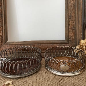 May include: Two antique silver-plated coasters with wooden bases. The coasters have a decorative wire design around the rim and a wooden base. The coasters are displayed on a burlap surface, with a vintage wooden frame and dried flowers in the background.