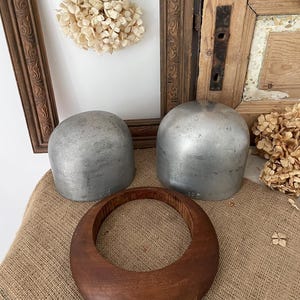 Könnte beinhalten: Zwei silberne Hutformen aus Metall und ein großer, runder Hutblock aus Holz liegen auf einer Jutefläche. Ein dekorativer Holzrahmen und Trockenblumen befinden sich im Hintergrund. Die Hutformen haben eine abgerundete, kuppelförmige Form.