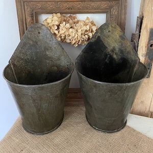 May include: Two antique metal wall planters with a weathered, aged appearance. The planters are a dark, muted grey colour and have a conical shape with a flat back and a small hole at the top for hanging. The planters are displayed on a burlap surface.