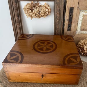 May include: A wooden box with a decorative inlaid design on the lid. The box is rectangular with a keyhole on the front. The lid features a central floral design and corner accents in a darker wood tone. Dried flowers and a vintage frame are in the background.