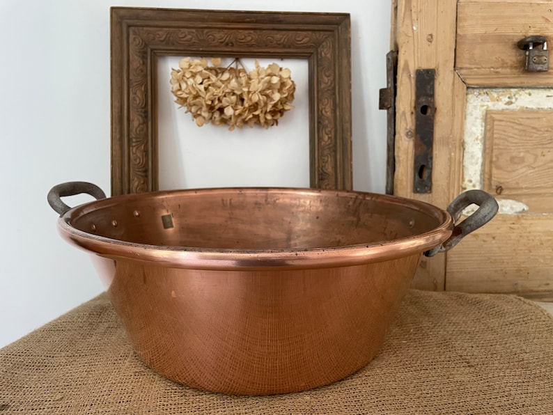 Puede incluir: Una olla grande de cobre con dos asas de hierro. La olla es redonda y tiene una superficie brillante y reflectante. Est&aacute; sentada sobre una superficie de arpillera marr&oacute;n.