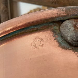 Puede incluir: Primer plano de una olla de cobre con un sello circular que dice "Fabrication Fran&ccedil;aise" en franc&eacute;s.