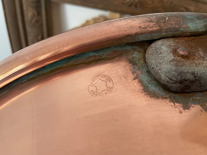 Puede incluir: Primer plano de una olla de cobre con un sello circular que dice "Fabrication Fran&ccedil;aise" en franc&eacute;s.