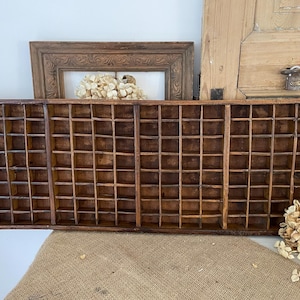 May include: A rectangular wooden printer's tray featuring a grid of numerous small, open compartments. The wood has a medium brown finish with visible grain and signs of wear. The tray is divided into four vertical sections by thicker dividers.
