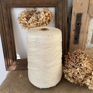 May include: A large spool of natural white yarn sits on a burlap surface. The spool is made of cardboard and has a wooden top. Dried hydrangeas are on either side of the spool.