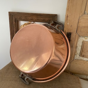 Puede incluir: Una olla grande de cobre con dos asas de hierro. La olla es brillante y tiene un aspecto desgastado y envejecido.