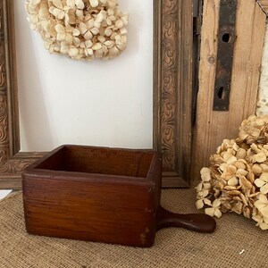 May include: A rectangular wooden box with a smooth, dark brown finish and a small, rounded handle extending from one side. The container features thick, solid walls and an open top, showcasing a simple, functional design with visible wood grain.