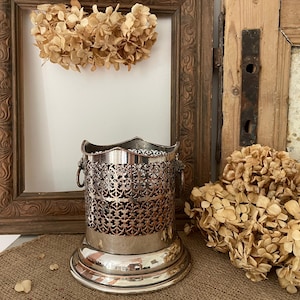 May include: A silver metal cup holder with a decorative openwork design and a scalloped rim. The cup holder has two small ring handles and sits on a round base. Dried flowers and a wooden picture frame are in the background.
