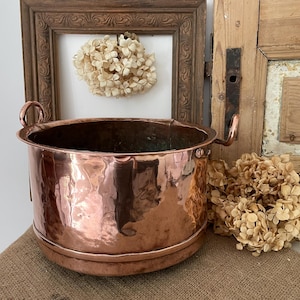 May include: A copper planter with a hammered finish and two curved handles. The round vessel has a shiny surface and a wide opening. The planter sits on a burlap surface, with dried flowers and a framed artwork in the background.